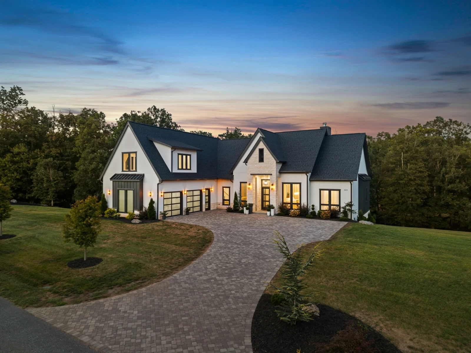 Twilight drone photo of a white farmhouse with a stone entry and a curved paver driveway